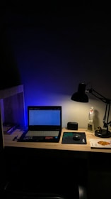 A dimly lit desk with a laptop displaying a webpage. The desk is organized with a few books on the side, a mouse resting on a mouse pad, and a lamp casting light onto the workspace. A figurine and a small speaker are also present, adding personal touches to the setup.