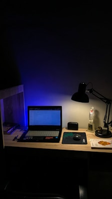 A dimly lit desk with a laptop displaying a webpage. The desk is organized with a few books on the side, a mouse resting on a mouse pad, and a lamp casting light onto the workspace. A figurine and a small speaker are also present, adding personal touches to the setup.