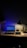 A dimly lit desk with a laptop displaying a webpage. The desk is organized with a few books on the side, a mouse resting on a mouse pad, and a lamp casting light onto the workspace. A figurine and a small speaker are also present, adding personal touches to the setup.