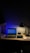 A dimly lit desk with a laptop displaying a webpage. The desk is organized with a few books on the side, a mouse resting on a mouse pad, and a lamp casting light onto the workspace. A figurine and a small speaker are also present, adding personal touches to the setup.