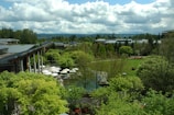 A lush, expansive garden landscape with a variety of green trees and shrubs, surrounding modern buildings with large glass windows and grey roofs. White umbrellas are visible over an outdoor seating area, along with well-maintained lawns and a few paved paths. The sky is partly cloudy, adding depth to the background with distant hills visible.