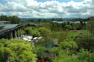 A lush, expansive garden landscape with a variety of green trees and shrubs, surrounding modern buildings with large glass windows and grey roofs. White umbrellas are visible over an outdoor seating area, along with well-maintained lawns and a few paved paths. The sky is partly cloudy, adding depth to the background with distant hills visible.