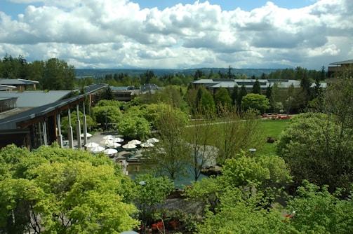 A lush, expansive garden landscape with a variety of green trees and shrubs, surrounding modern buildings with large glass windows and grey roofs. White umbrellas are visible over an outdoor seating area, along with well-maintained lawns and a few paved paths. The sky is partly cloudy, adding depth to the background with distant hills visible.