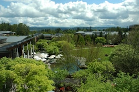 A lush, expansive garden landscape with a variety of green trees and shrubs, surrounding modern buildings with large glass windows and grey roofs. White umbrellas are visible over an outdoor seating area, along with well-maintained lawns and a few paved paths. The sky is partly cloudy, adding depth to the background with distant hills visible.