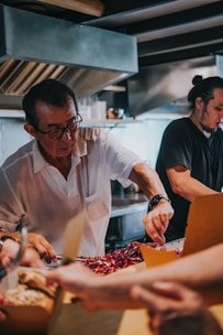 A busy kitchen scene with two individuals preparing food. The person wearing a white shirt is concentrating on arranging ingredients, possibly involving sliced vegetables. The kitchen environment appears industrial, equipped with stainless steel appliances and an extractor hood.