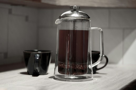 A glass French press filled with dark liquid stands on a light wooden surface, accompanied by two black ceramic mugs. The background consists of tiled walls, giving a clean and modern kitchen atmosphere.