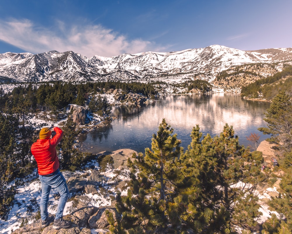 person in red jacket standing on rocky mountain near lake during daytime