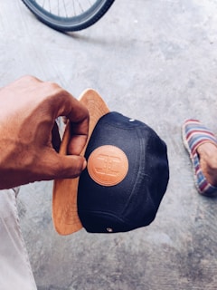 A hand holding a black cap with a brown leather patch featuring a logo. The cap has a wooden visor that matches the patch. In the background, part of a bicycle wheel is visible along with a foot wearing a striped slip-on shoe on a textured concrete surface.