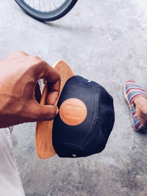 A hand holding a black cap with a brown leather patch featuring a logo. The cap has a wooden visor that matches the patch. In the background, part of a bicycle wheel is visible along with a foot wearing a striped slip-on shoe on a textured concrete surface.