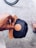 A hand holding a black cap with a brown leather patch featuring a logo. The cap has a wooden visor that matches the patch. In the background, part of a bicycle wheel is visible along with a foot wearing a striped slip-on shoe on a textured concrete surface.