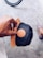 A hand holding a black cap with a brown leather patch featuring a logo. The cap has a wooden visor that matches the patch. In the background, part of a bicycle wheel is visible along with a foot wearing a striped slip-on shoe on a textured concrete surface.