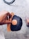 A hand holding a black cap with a brown leather patch featuring a logo. The cap has a wooden visor that matches the patch. In the background, part of a bicycle wheel is visible along with a foot wearing a striped slip-on shoe on a textured concrete surface.