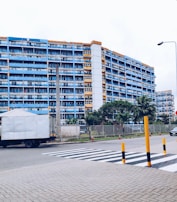 A multi-story residential building with blue and orange accents is situated behind a street with a zebra crossing. The foreground shows a paved area and a large delivery truck parked on the side of the road. There are several tall palm trees and green shrubs lining the building, and orange and black bollards are placed near the crossing. A single streetlamp is visible, and a car is seen driving in the background.