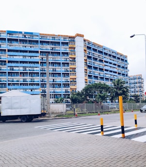A multi-story residential building with blue and orange accents is situated behind a street with a zebra crossing. The foreground shows a paved area and a large delivery truck parked on the side of the road. There are several tall palm trees and green shrubs lining the building, and orange and black bollards are placed near the crossing. A single streetlamp is visible, and a car is seen driving in the background.