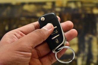 A hand is holding a car key fob, which features buttons for locking and unlocking the vehicle, and possibly an alarm function. The background is blurred and consists of various shades of brown and yellow.