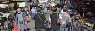 A bustling outdoor market scene with numerous people walking and shopping. Many are wearing masks, indicating an awareness of health precautions. Stalls line both sides of the pathway, offering a variety of goods such as fruits, vegetables, and other market items. The atmosphere is busy, with people engaged in different activities like purchasing and carrying bags.