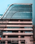 A modern multi-story office building with a distinctive wavy architectural design. The facade is made predominantly of glass and metal, with an array of vertical and horizontal lines creating a grid-like pattern. There are signs for 'National Insurance' and 'Parking 24x7' visible in multiple languages. The structure appears robust and industrial.