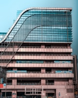A modern multi-story office building with a distinctive wavy architectural design. The facade is made predominantly of glass and metal, with an array of vertical and horizontal lines creating a grid-like pattern. There are signs for 'National Insurance' and 'Parking 24x7' visible in multiple languages. The structure appears robust and industrial.