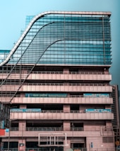 A modern multi-story office building with a distinctive wavy architectural design. The facade is made predominantly of glass and metal, with an array of vertical and horizontal lines creating a grid-like pattern. There are signs for 'National Insurance' and 'Parking 24x7' visible in multiple languages. The structure appears robust and industrial.