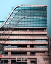 A modern multi-story office building with a distinctive wavy architectural design. The facade is made predominantly of glass and metal, with an array of vertical and horizontal lines creating a grid-like pattern. There are signs for 'National Insurance' and 'Parking 24x7' visible in multiple languages. The structure appears robust and industrial.