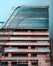 A modern multi-story office building with a distinctive wavy architectural design. The facade is made predominantly of glass and metal, with an array of vertical and horizontal lines creating a grid-like pattern. There are signs for 'National Insurance' and 'Parking 24x7' visible in multiple languages. The structure appears robust and industrial.