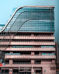 A modern multi-story office building with a distinctive wavy architectural design. The facade is made predominantly of glass and metal, with an array of vertical and horizontal lines creating a grid-like pattern. There are signs for 'National Insurance' and 'Parking 24x7' visible in multiple languages. The structure appears robust and industrial.