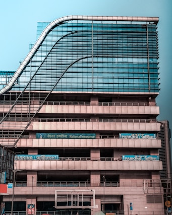 A modern multi-story office building with a distinctive wavy architectural design. The facade is made predominantly of glass and metal, with an array of vertical and horizontal lines creating a grid-like pattern. There are signs for 'National Insurance' and 'Parking 24x7' visible in multiple languages. The structure appears robust and industrial.
