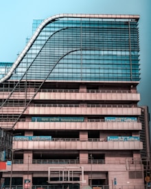 A modern multi-story office building with a distinctive wavy architectural design. The facade is made predominantly of glass and metal, with an array of vertical and horizontal lines creating a grid-like pattern. There are signs for 'National Insurance' and 'Parking 24x7' visible in multiple languages. The structure appears robust and industrial.