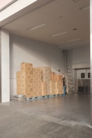 A stack of cardboard boxes is neatly arranged on pallets in a warehouse or storage area. The space is well-lit with overhead fluorescent lights, and there's a metal ladder leaning against the wall. The floor is concrete, and large double doors are partially visible.