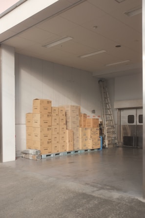 A stack of cardboard boxes is neatly arranged on pallets in a warehouse or storage area. The space is well-lit with overhead fluorescent lights, and there's a metal ladder leaning against the wall. The floor is concrete, and large double doors are partially visible.