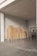 A stack of cardboard boxes is neatly arranged on pallets in a warehouse or storage area. The space is well-lit with overhead fluorescent lights, and there's a metal ladder leaning against the wall. The floor is concrete, and large double doors are partially visible.