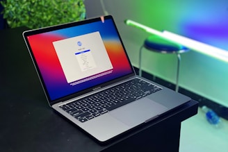 A sleek laptop with a colorful screen displaying a language selection window. The device sits on a dark surface against a contrasting backdrop of vivid lights and softly focused greenery, creating a modern and high-tech atmosphere.