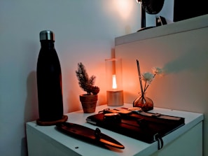 A neatly arranged tabletop featuring a black water bottle on a coaster, a small potted pine plant, a cylindrical table lamp emitting a warm glow, and a round glass vase with reeds and white flowers. Additionally, a closed notebook and a pen can be seen in the scene, creating a cozy, minimalist aesthetic.