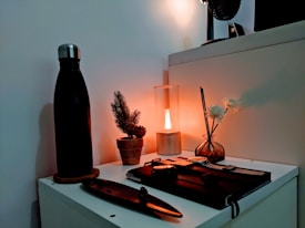 A neatly arranged tabletop featuring a black water bottle on a coaster, a small potted pine plant, a cylindrical table lamp emitting a warm glow, and a round glass vase with reeds and white flowers. Additionally, a closed notebook and a pen can be seen in the scene, creating a cozy, minimalist aesthetic.