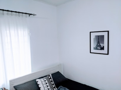 A minimalist bedroom with white walls, featuring a black and white photograph of the Eiffel Tower in a black frame. The bed is against the wall with a monochrome geometric patterned pillow and dark sheets. Sheer white curtains partially cover a window, allowing natural light to enter.