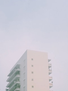 A minimalist, modern apartment building with a pale exterior set against a muted sky. The structure features simple geometric lines, balconies, and small windows embedded in its facade.
