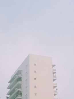 A minimalist, modern apartment building with a pale exterior set against a muted sky. The structure features simple geometric lines, balconies, and small windows embedded in its facade.