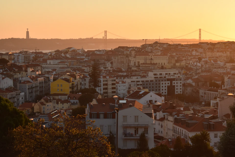 Lisbon's rooftops glowing orange at sunset, with the 25 de Abril Bridge and the Cristo Rei statue on the far bank of the Tagus in the haze