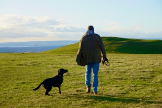 man in blue jacket standing beside black labrador retriever on green grass field during daytime