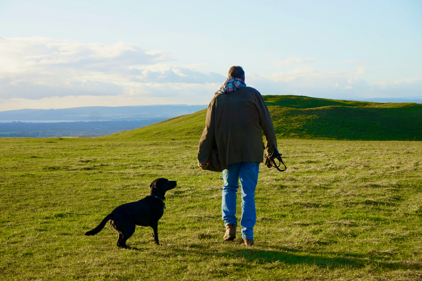A man walking outdoors across an open grassy field with his black dog close at his side in soft daylight