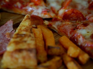 Slices of pizza with pepperoni and melted cheese are visible alongside crispy sweet potato fries. The thin crust pizza features a generous serving of toppings, including pepperoni and ham.