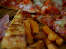 Slices of pizza with pepperoni and melted cheese are visible alongside crispy sweet potato fries. The thin crust pizza features a generous serving of toppings, including pepperoni and ham.