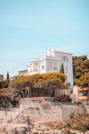 A grand white house perched on a rocky cliff, surrounded by lush greenery and tall pine trees. The house features a classical architectural style with a flat roof and decorative railings. It overlooks a serene seaside landscape with clear sky above.