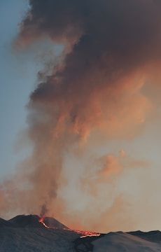 A dark volcanic eruption with thick smoke and ash clouds rising into the sky. The scene captures fiery lava flowing down the volcano's slopes against a backdrop of grey and orange clouds.