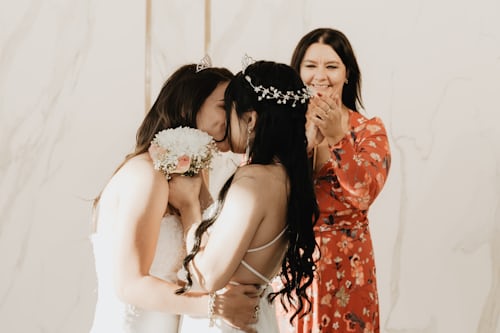 Deux femmes en robes de mariées s'embrassent devant une femme en robe à fleurs qui applaudit