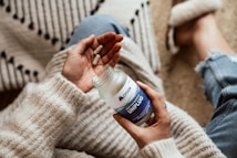 A person is sitting on a soft surface, dressed in a cozy knitted sweater and blue jeans, holding a container labeled 'Unplug' with a couple of capsules. The scene suggests a relaxing environment with a focus on self-care or wellness.