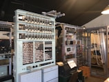 A vintage computing machine with numerous cables, dials, and switches in a room filled with historic equipment. The setup includes large metallic frames holding intricate components like relays and wires. There’s also an old-type computer terminal and other associated apparatus.