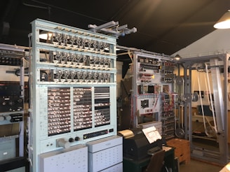 A vintage computing machine with numerous cables, dials, and switches in a room filled with historic equipment. The setup includes large metallic frames holding intricate components like relays and wires. There’s also an old-type computer terminal and other associated apparatus.