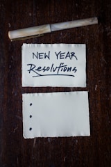 Two pieces of torn paper rest on a dark wooden surface. The top paper has 'NEW YEAR Resolutions' written in bold, black lettering. A pen is placed above the papers. The bottom paper has three bullet points but is otherwise blank.