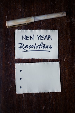 Two pieces of torn paper rest on a dark wooden surface. The top paper has 'NEW YEAR Resolutions' written in bold, black lettering. A pen is placed above the papers. The bottom paper has three bullet points but is otherwise blank.